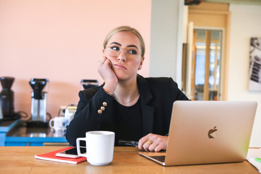 Woman in a black blazer sitting at a table with a laptop and coffee mug, looking bored or contemplative in a modern office setting.