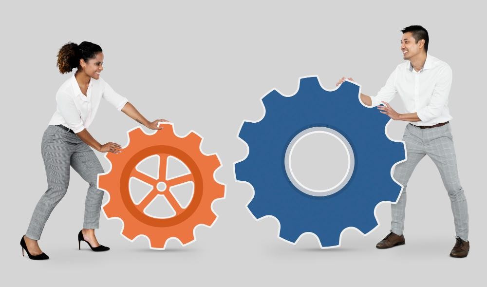 Smiling man and woman pushing large interlocking gears together, symbolizing teamwork and collaboration.