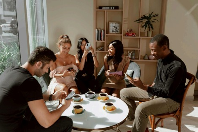 Group of young adults sitting at a coffee shop, each using smartphones and engaging with social media apps - representing Snapchat’s ideal target audience under 30.