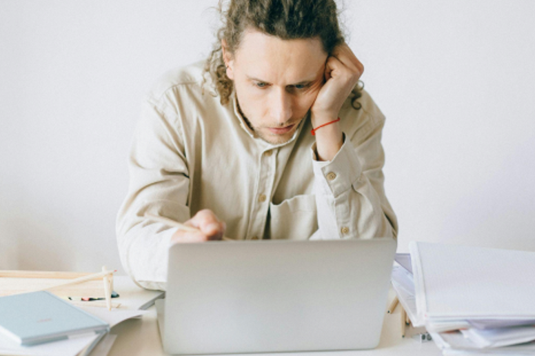 Overworked office worker surrounded by papers and sticky notes, staring at multiple computer screens with a stressed expression.